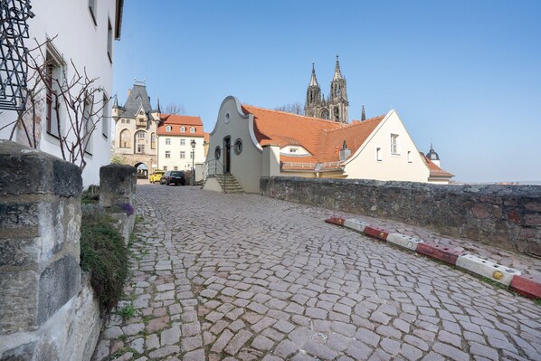 Une rue pavée menant à un bâtiment historique, doté d’un toit recouvert de tuiles rouges, et une église avec deux flèches jumelles en arrière-plan.