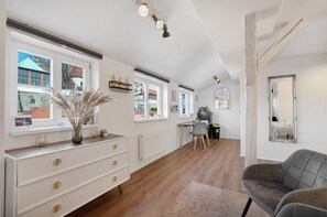 Une pièce spacieuse avec une grande fenêtre, un plancher en bois, une commode blanche, une chaise grise et un miroir au mur.