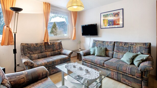 Ein Wohnzimmer mit gemusterten Sofas, einem Glastisch, einer Stehlampe, einem Fernseher und einem fenster mit Vorhängen.