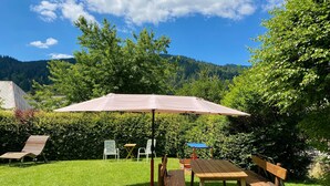 Ein Picknickplatz mit Tisch, Bänken und Sonnenschirm in einer grünen Wiese mit Bäumen und einem Berg im Hintergrund.