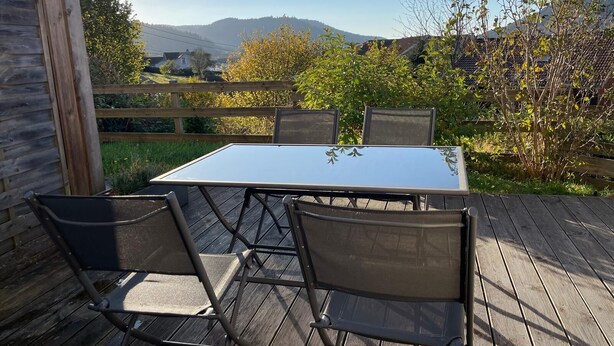 Un patio al aire libre con una mesa y cuatro sillas, rodeado por una terraza de madera y vistas a un paisaje montañoso.