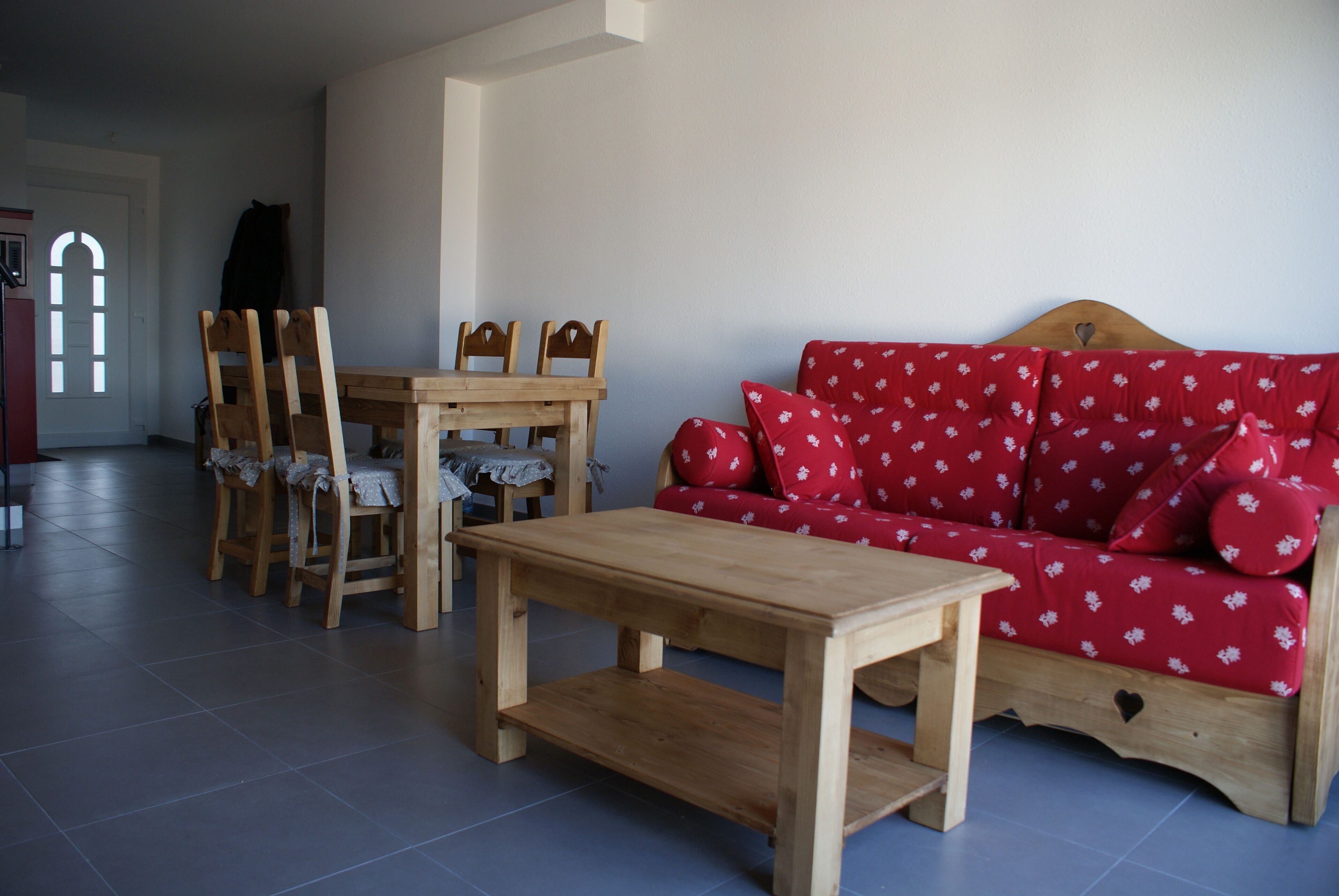 A living room with a wooden table and chairs, a red sofa with floral cushions, and a wooden bench.