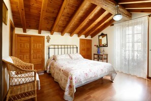 Une chambre avec un plafond en bois, un lit recouvert d’une couvre-lit à motifs floraux, une chaise en osier et une armoire en bois.