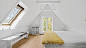 A high-ceilinged, white-walled room with a sloped ceiling, a skylight, a large window, a bed, a bedside table, and a bench. - Cozy home in Aakirkeby with WiFi (Aakirkeby)