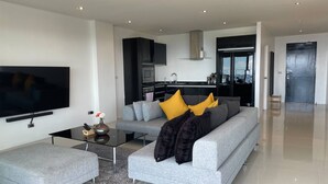 A modern living room with a grey sofa, ottoman, and a flat-screen TV. - A26 - 2 Bdr Condo, Stunning Sea View, Karon Beach (Karon)