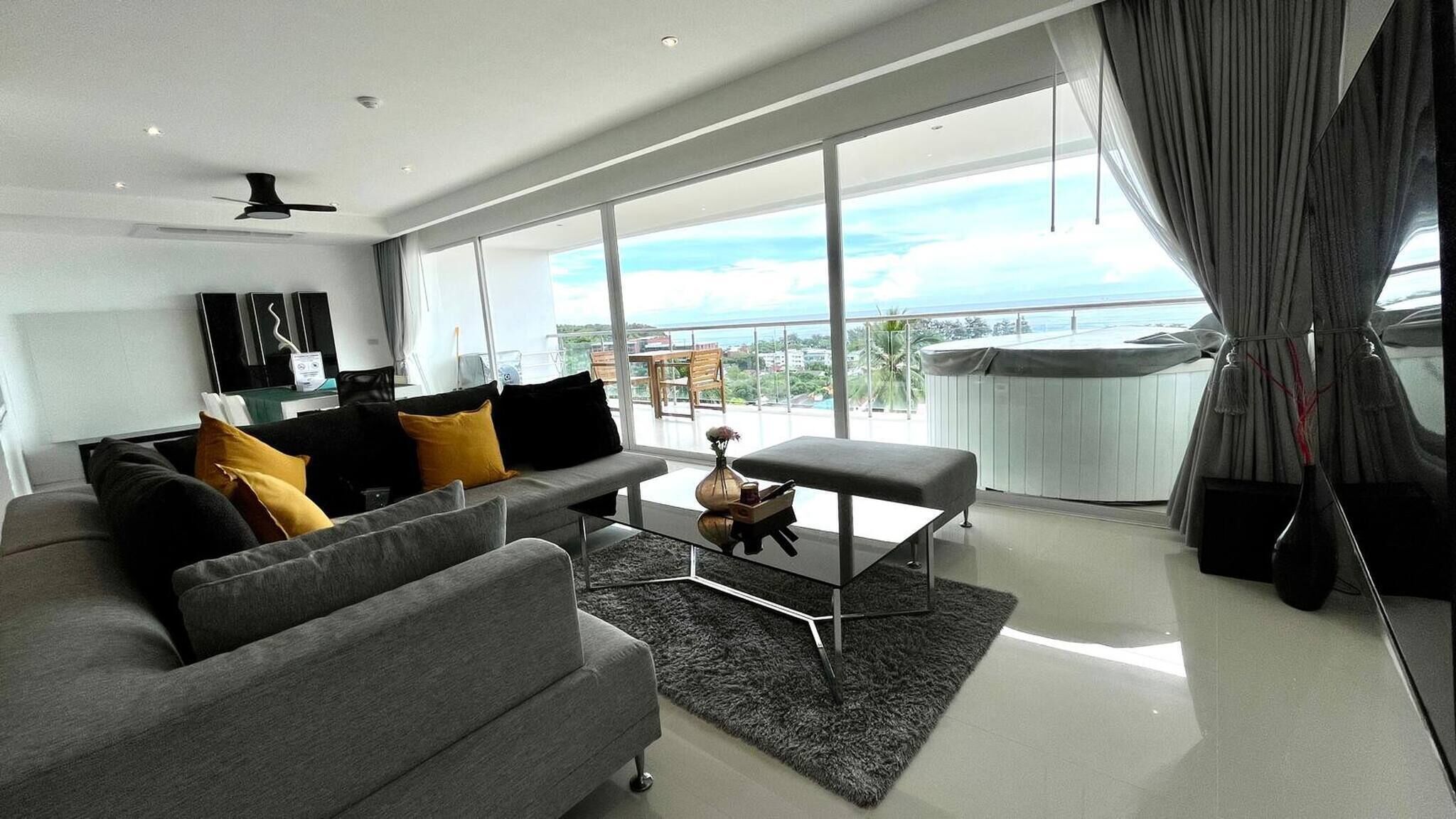 A modern living room with a grey sofa, black and yellow cushions, a glass coffee table, and a view of the sea through large windows.