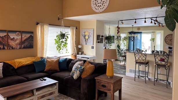 Una sala de estar acogedora con un sofá modular, una mesa de centro y una mesita de noche con lámpara. Se observa un comedor con sillas y una mesa al fondo.
