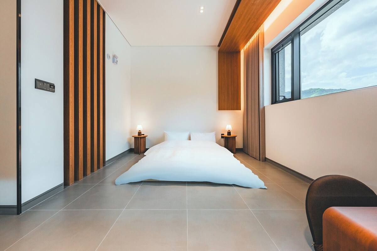 A modern hotel room with a large bed, wooden paneling on the walls, and a view of the outdoors through a large window.