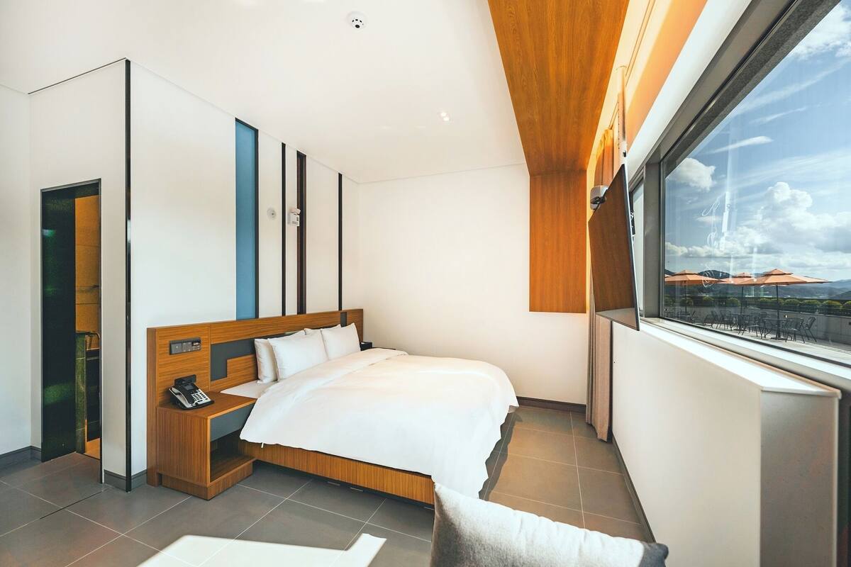 A modern hotel room with a large bed, a bedside table with a phone, a window with a view of the outdoors, and a wooden ceiling.