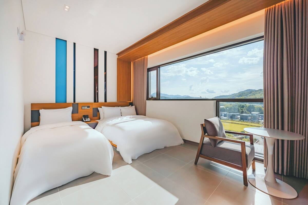 A modern hotel room with two beds, a small table, and a view of the outdoors.