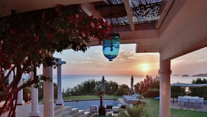 Un espacio exterior cubierto con piscina, columnas blancas y vistas al mar al atardecer.