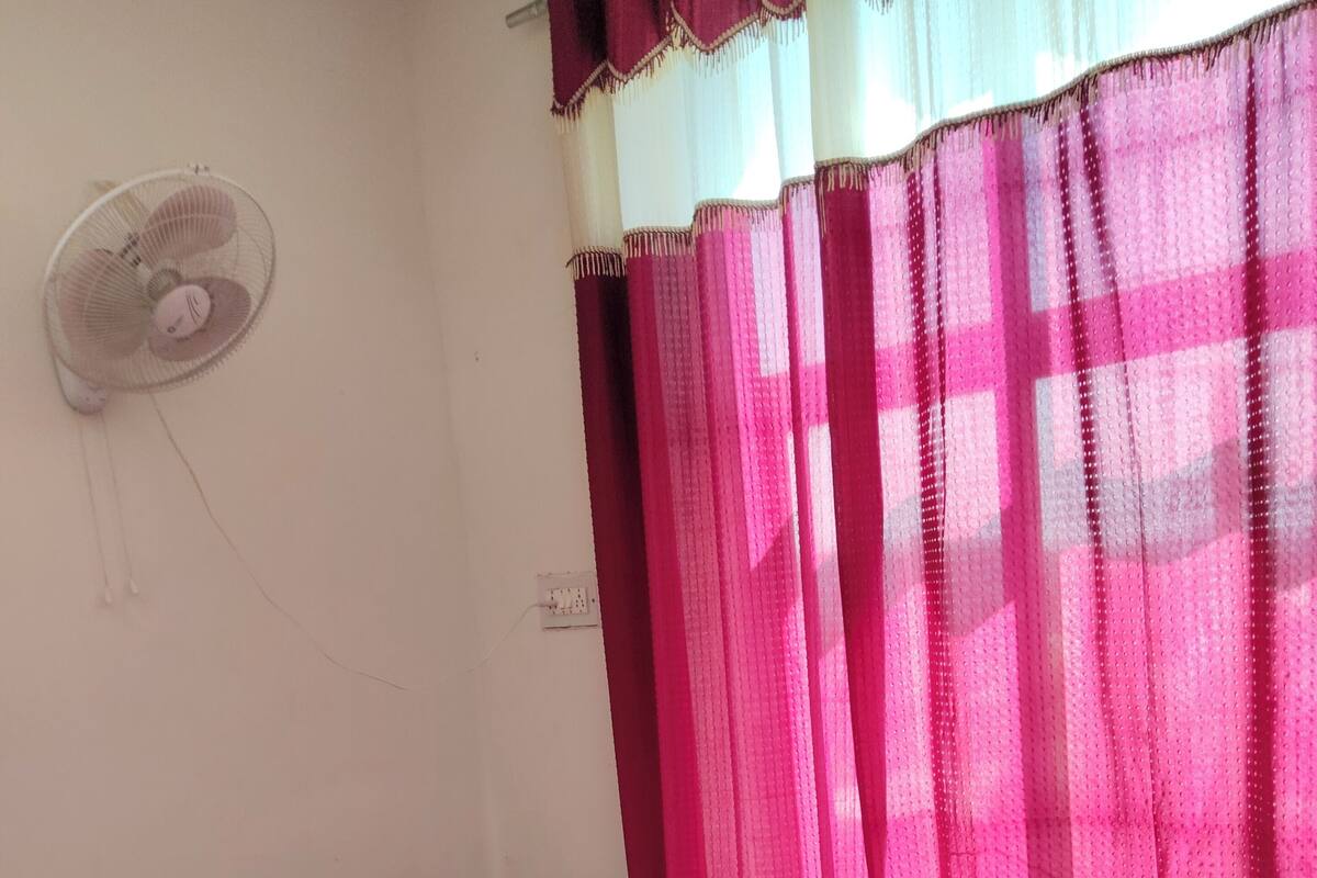 A room with a bed, a chair, a standing fan, and a window with pink curtains.