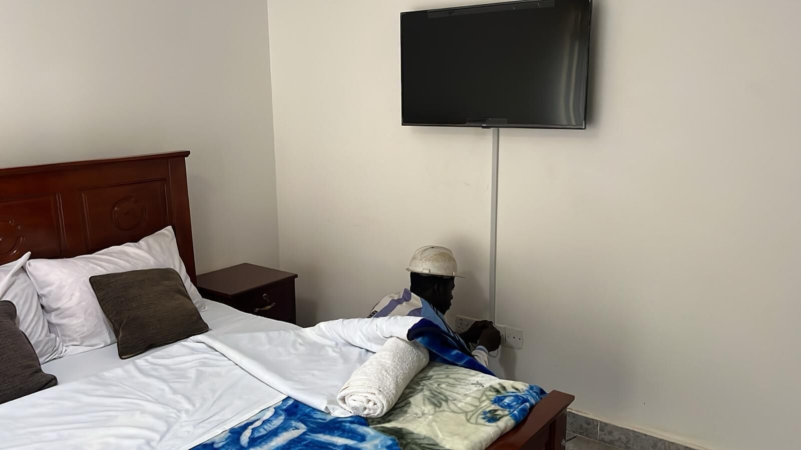Une chambre d’hôtel avec un lit, une table de chevet en bois, un téléviseur fixé au mur et un mannequin vêtu d’une tenue de travail.