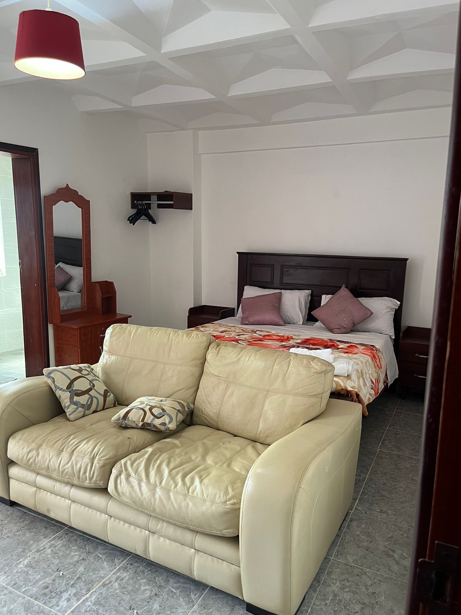 A room with a beige leather sofa, a wooden headboard bed, a mirror, and a red pendant light.
