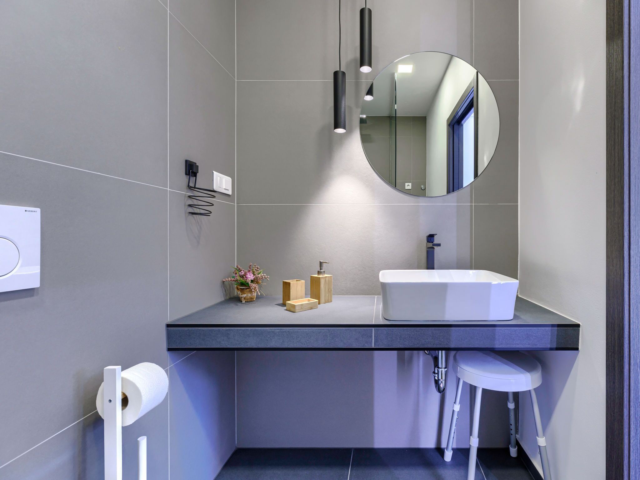 A modern bathroom with a grey tiled wall, a white sink, a round mirror, and a toilet with a white seat.