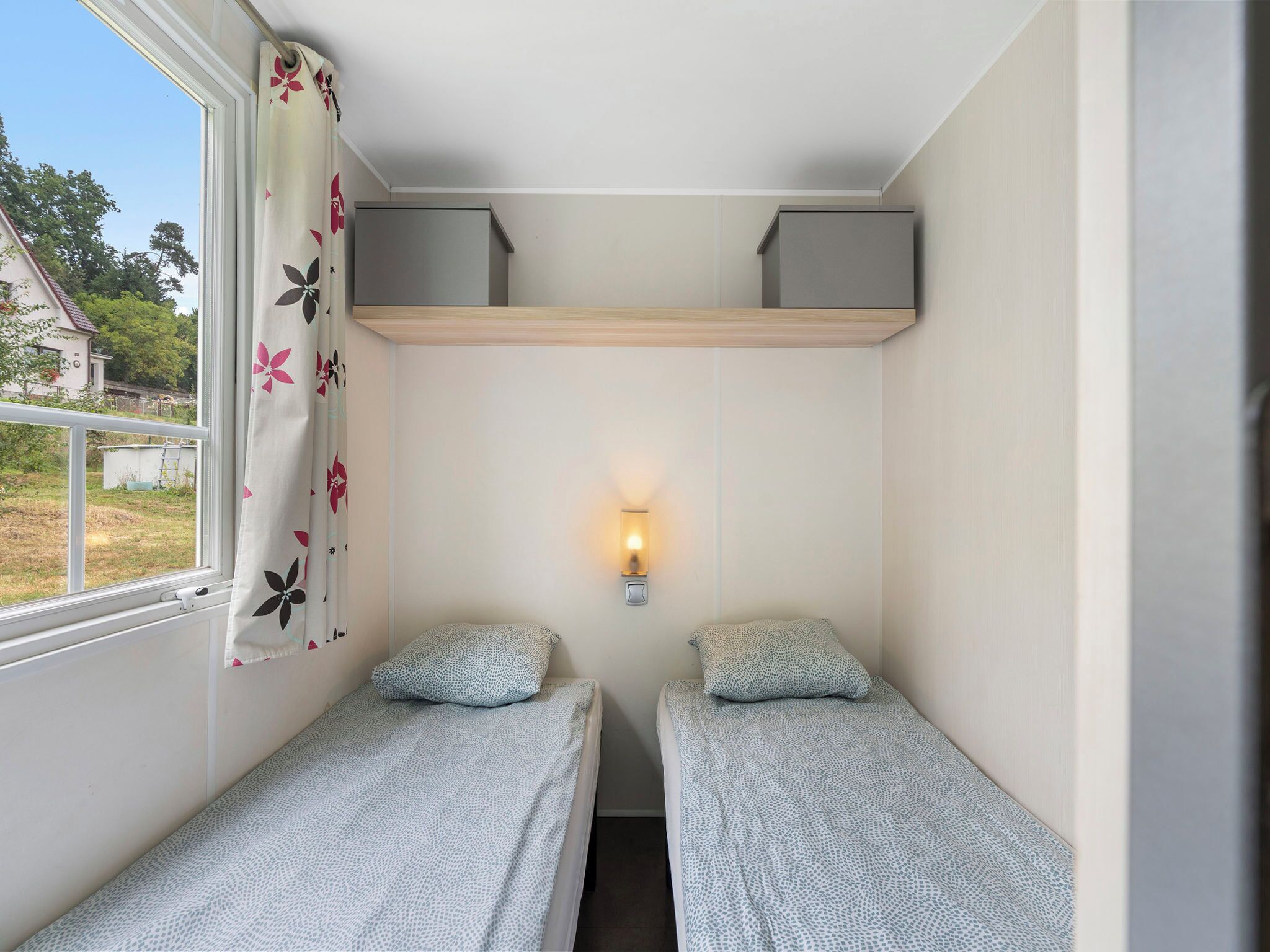 Een kleine, eenpersoons accommodatie met twee eenpersoonsbedden, een raam met bloemeninrichting, een wandlamp en uitzicht op buiten.