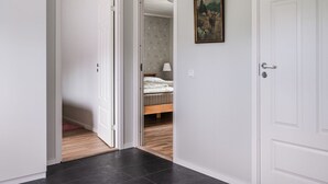 Un pasillo con dos puertas blancas, una de ellas da a un dormitorio con cama y mesita de noche, y un cuadro en la pared.