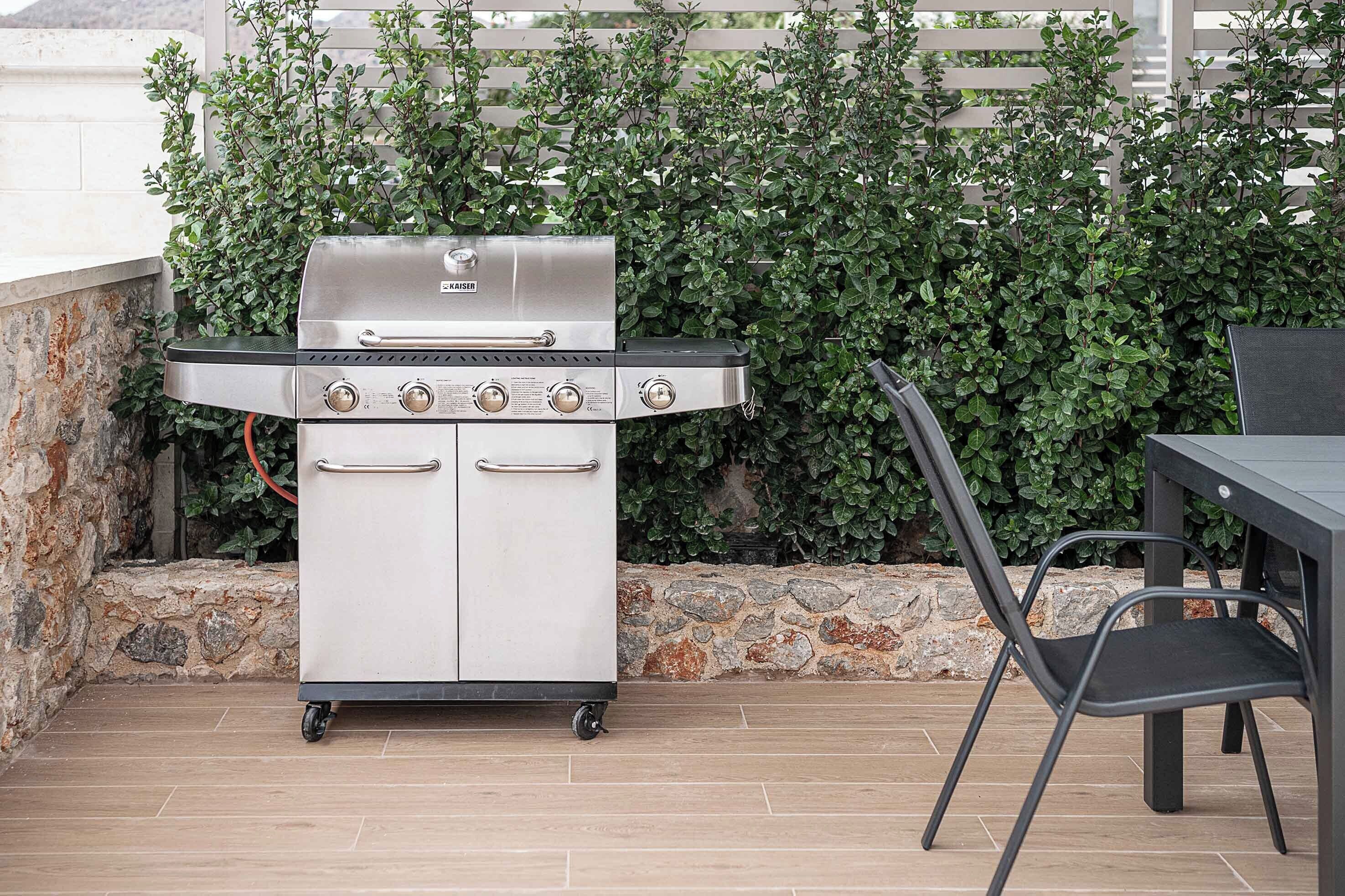 A portable stainless steel grill with four burners, a side cabinet, and a foldable chair, set against a stone wall and green foliage.