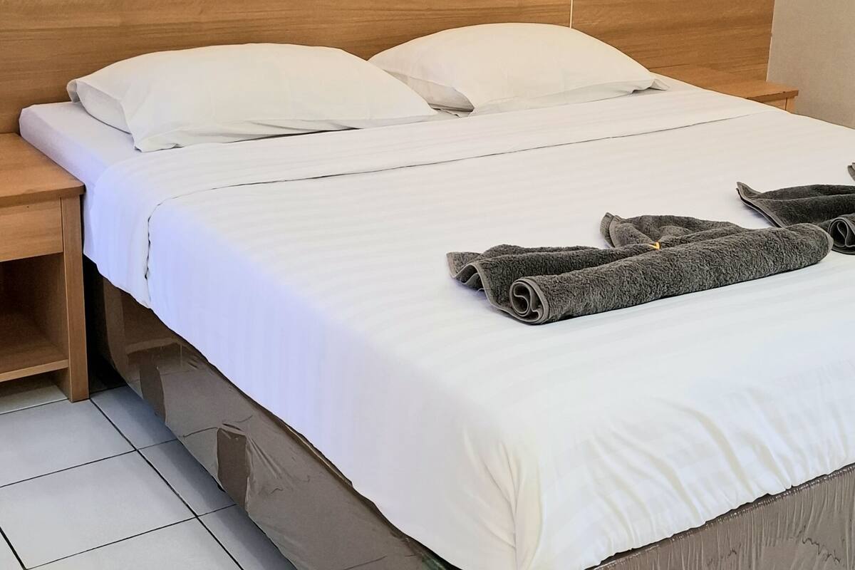 Una cama bien hecha con sábanas blancas, un cabecero de madera y una mesita de noche con una lámpara.