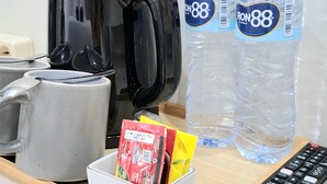 A kitchen area with a coffee maker, two water bottles, a remote control, and a box of snacks. - Royal Kencana Hotel (Jepara)