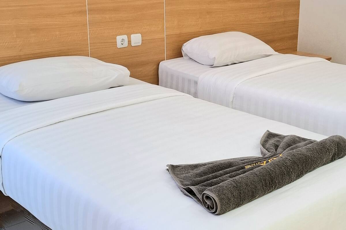 Una habitación de hotel con dos camas, cada una con ropa de cama y almohadas blancas. Una toalla enrollada está colocada en la cama en primer plano.