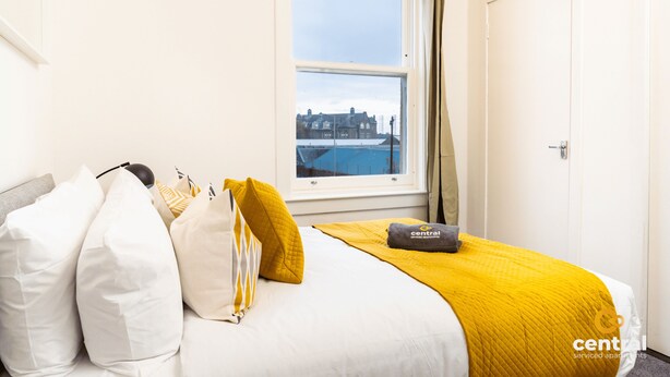 A hotel room with a bed, pillows, a blanket, and a view of a building through a window. - Court Street by Central Serviced Apartments (Dundee)