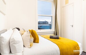 A hotel room with a bed, pillows, a blanket, and a view of a building through a window.
