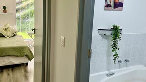 A bedroom with a bed, a window with blinds, and a door leading to a bathroom with a bathtub and a towel rack. - Charming 2-bedroom apartment in the beautifully designed Gary Player Golf Estate (Plettenberg Bay)