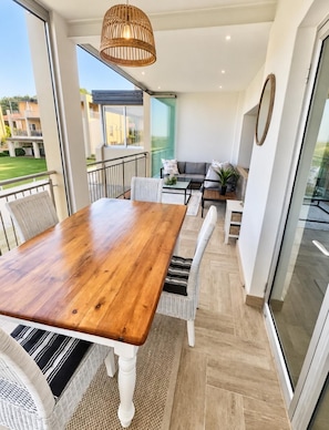 Een eetgedeelte met een houten tafel en witte stoelen, een hanglamp van wilg en een uitzicht op het buitenbalkon.