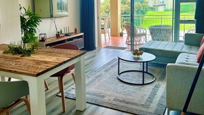 A modern living room with a wooden dining table, a sofa, and a TV mounted on the wall. - Charming 2-bedroom apartment in the beautifully designed Gary Player Golf Estate (Plettenberg Bay)