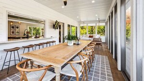 Une vaste terrasse couverte, dotée d’une longue table et de chaises en bois, d’un ventilateur de plafond et de grandes fenêtres offrant une vue sur la verdure.