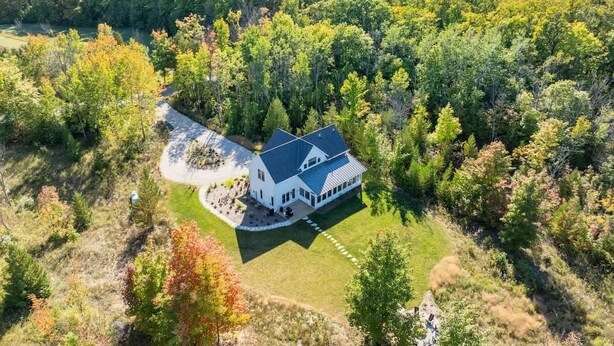 Une vue aérienne d’une grande maison blanche, dotée d’un toit foncé, entourée d’une forêt dense et avec une étendue d’eau en arrière-plan.