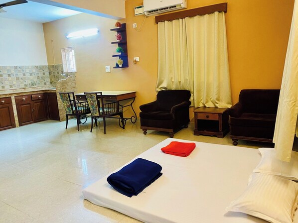 A hotel room with a bed, two armchairs, a small table, a desk, a chair, a window with curtains, and a kitchenette with cabinets and a tiled backsplash. - Nasya Beachside Cottages (Benaulim)