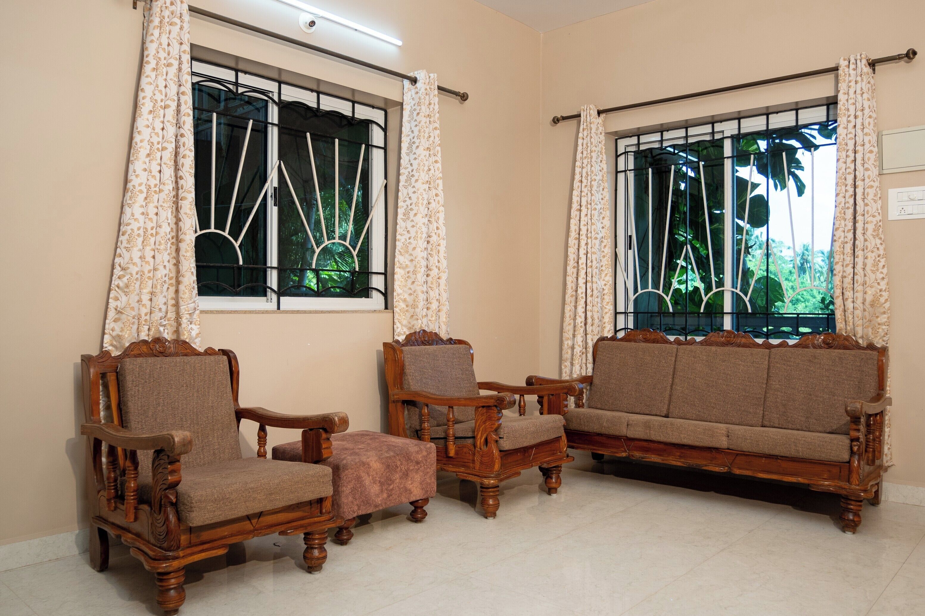 Una habitación con dos sofás de madera, dos sillones y un pequeño otoman. Hay dos ventanas con cortinas, una de ellas ofrece vistas a zonas verdes.