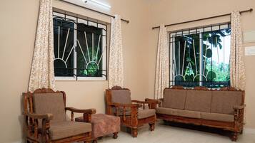 Una habitación con dos sofás de madera, dos sillones y un pequeño otoman. Hay dos ventanas con cortinas, una de ellas ofrece vistas a zonas verdes.