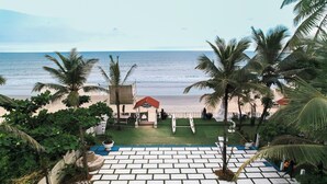 A beach view with palm trees, a white tiled area, and a small structure with a red roof. - Nasya Beachside Cottages (Benaulim)