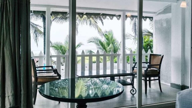 A room with a glass table and chairs, large sliding glass doors leading to a balcony with a white fence and palm trees. - Nasya Beachside Cottages (Benaulim)