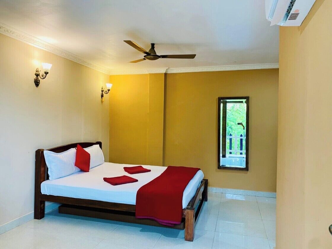 A single bed with a wooden frame, white bedding, and red accents. A ceiling fan is visible, and a window with a view of greenery is present.