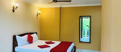 A single bed with a wooden frame, white bedding, and red accents. A ceiling fan is visible, and a window with a view of greenery is present.