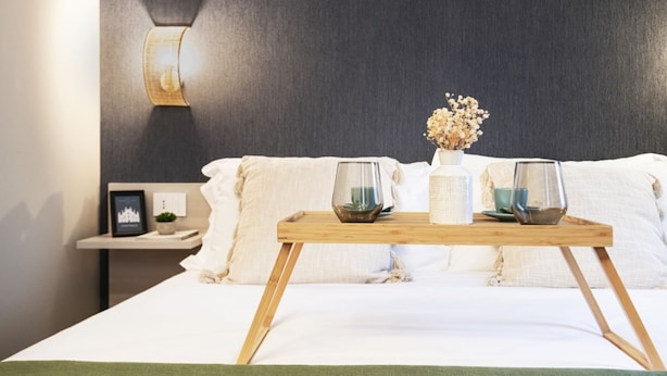 A neatly made bed with a wooden tray holding two glasses and a vase with dried flowers. A wall-mounted light fixture and a small bedside table with a picture frame and a plant are visible. - Easylife-Sophisticated  with Jacuzzi in Centrale (Milano)
