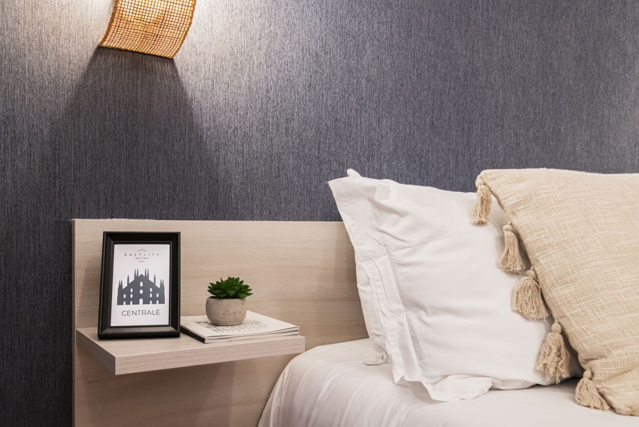 A neatly arranged hotel room with a bed, a bedside table with a framed picture, a small potted plant, and a wall-mounted light fixture.