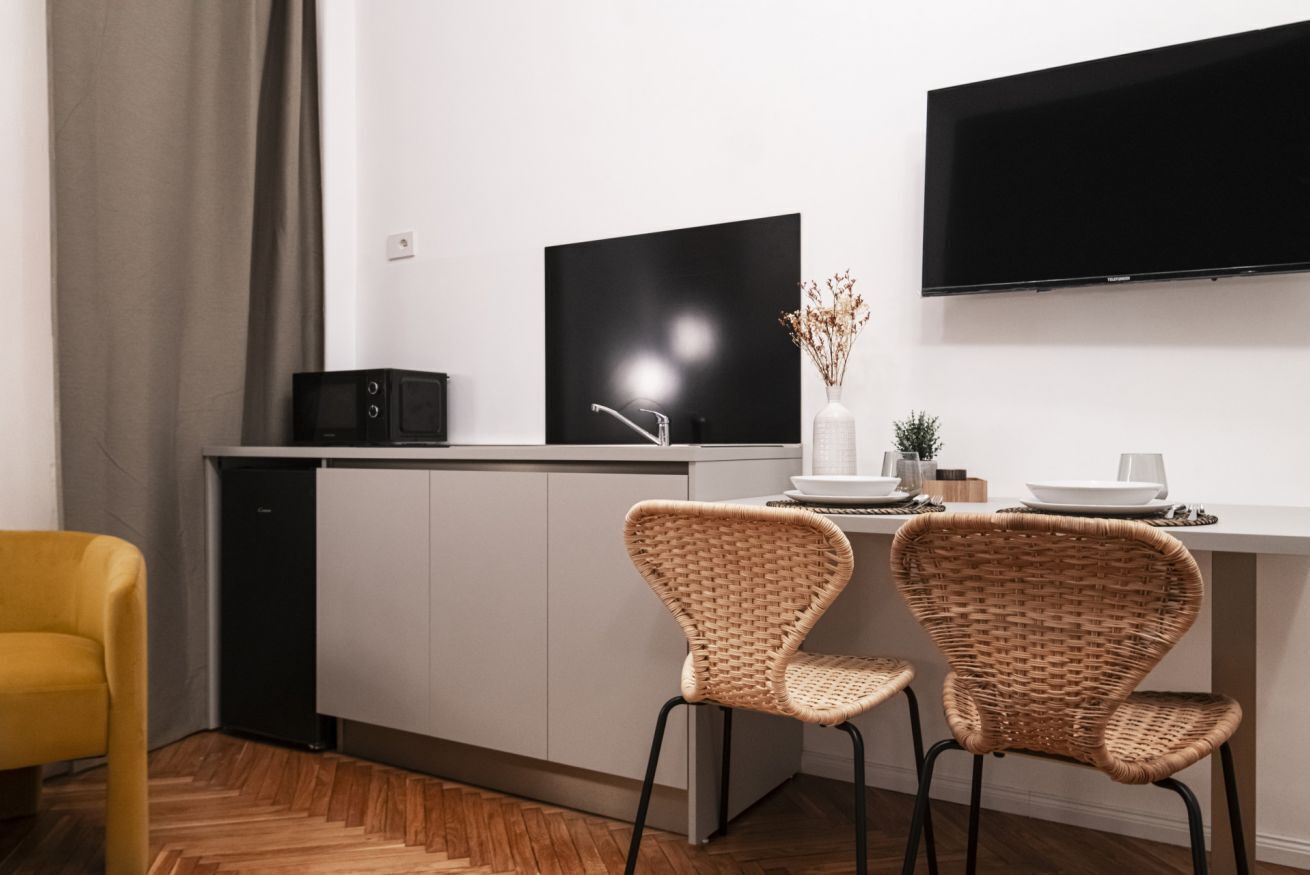 A modern kitchen with a flat-screen TV, two wicker chairs, a yellow chair, and a dining table set with plates and glasses.