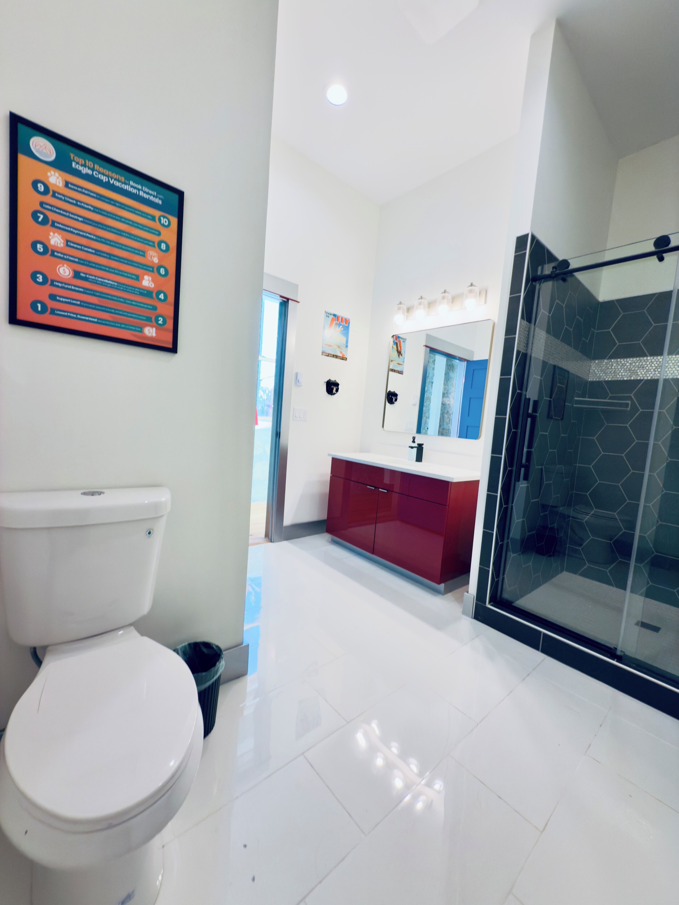 Un baño moderno con inodoro blanco, tocador rojo, cabina de ducha con mampara de vidrio y un cartel en la pared.