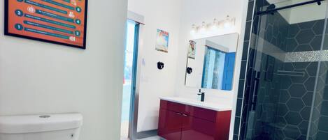 Un baño moderno con inodoro blanco, tocador rojo, cabina de ducha con mampara de vidrio y un cartel en la pared.