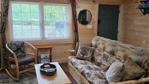 A cozy cabin interior with a wooden floor, a sofa with animal print cushions, a rocking chair, a table with a guitar, and a window with curtains. - Stone Haven Retreat by Beyond Expectations: Dog Friendly w/ Hot Tub & Fireplace (Sevierville)