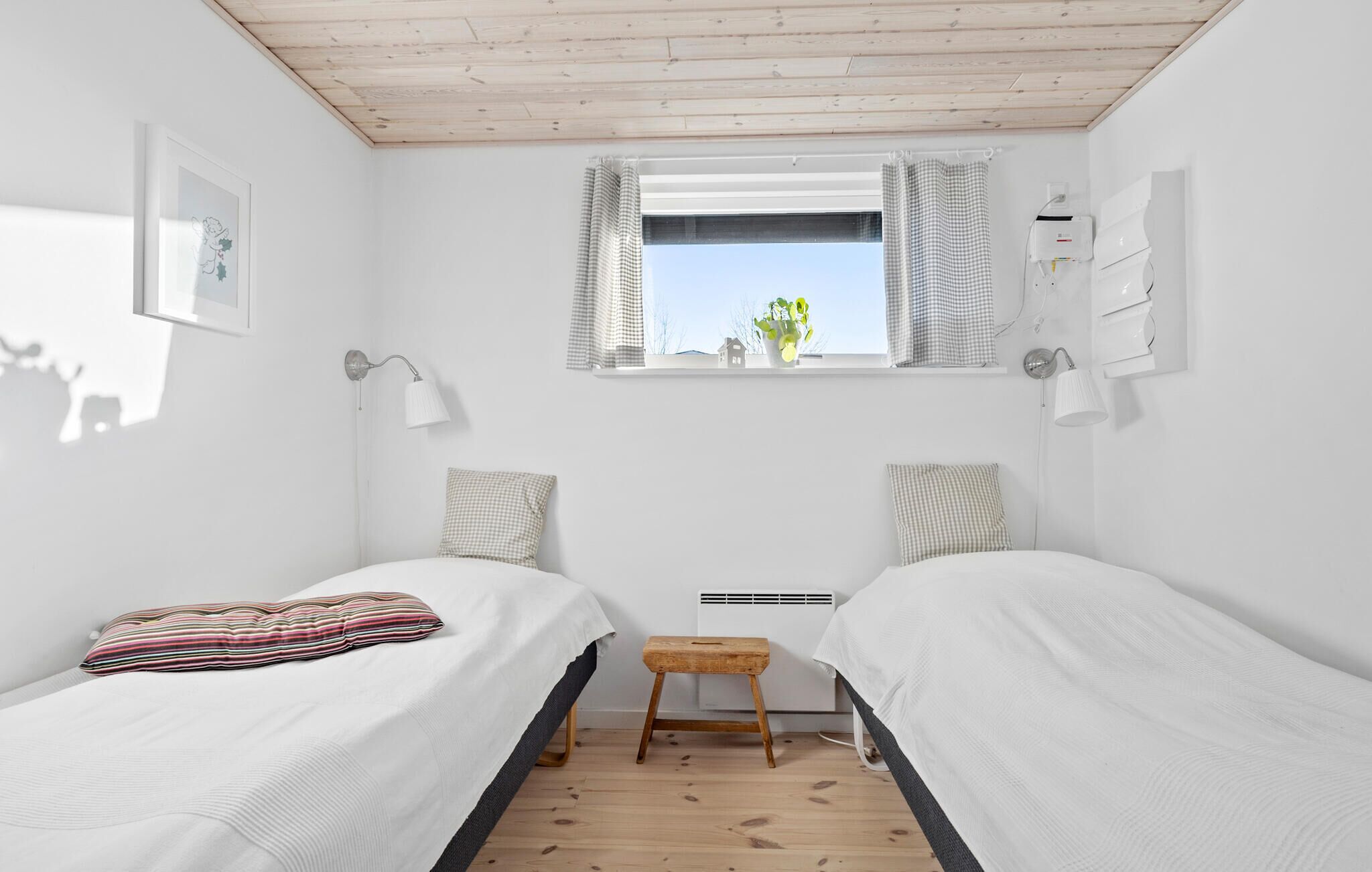 A bedroom with two single beds, a window with curtains, a small stool, and a wall-mounted heater.