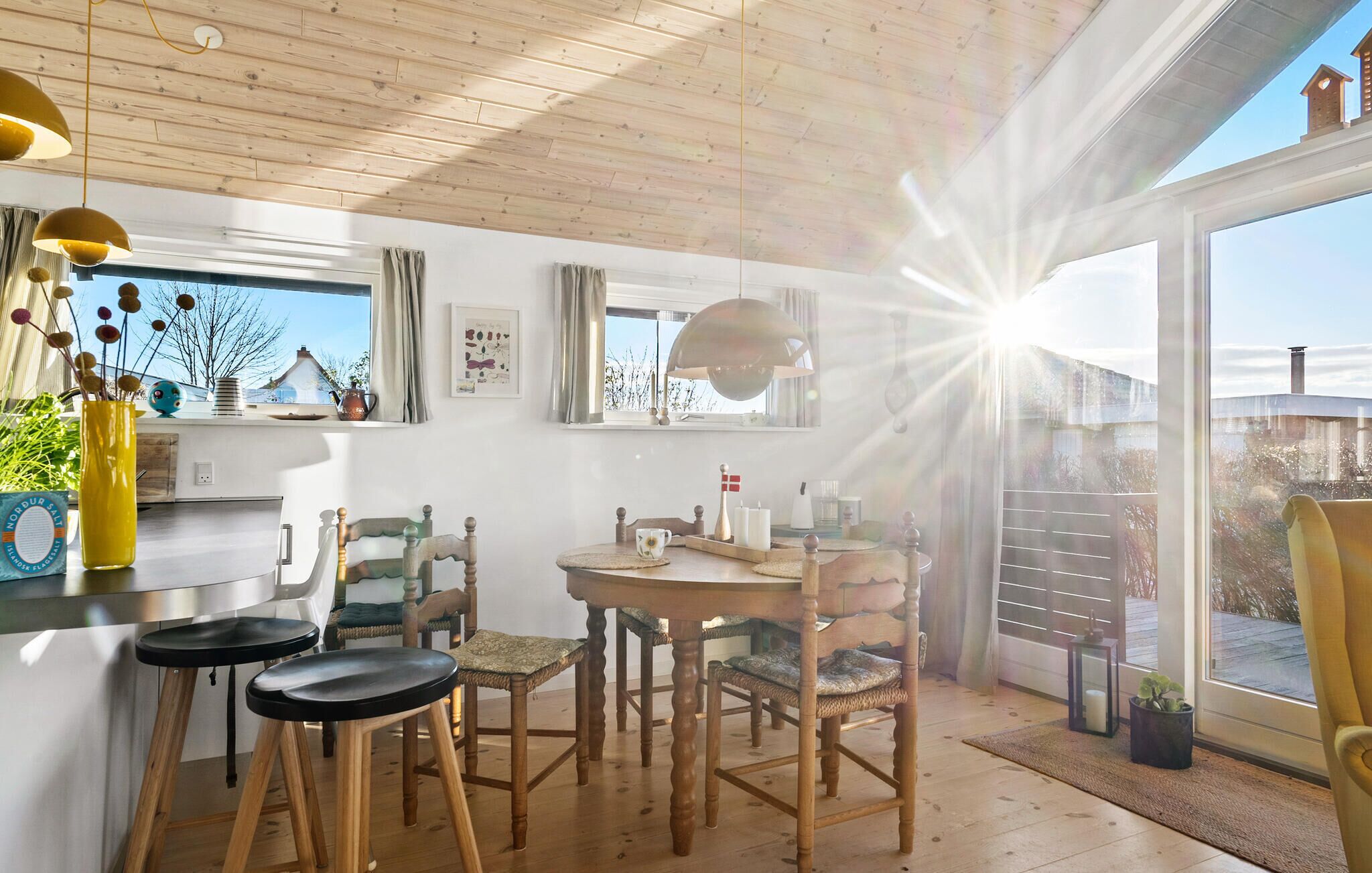 A cozy dining area with wooden furniture, a round table, and chairs. There is a window with curtains, a hanging light fixture, and a view of the outdoors.