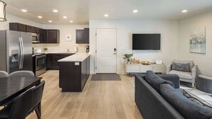 A modern kitchen with dark cabinets, a refrigerator, microwave, and oven. There is a dining area with a table and chairs, and a living room with a gray sofa and a TV on the wall. - 6G Moab RedCliff Condo, POOL & HOT TUB (Moab)