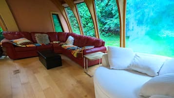Uma sala de estar espaçosa com um sofá vermelho, um sofá branco e uma mesa de centro de madeira. Grandes janelas oferecem vista para o verde lá fora.