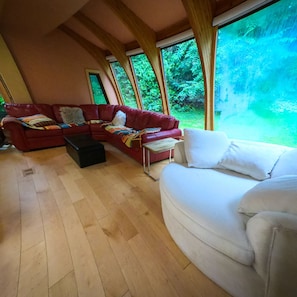 A spacious living room with a red sofa, a white sofa, and a wooden coffee table. Large windows provide a view of the greenery outside.